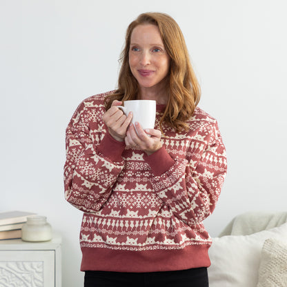A woman wearing a rust red and cream Fair Isle patterned sweater with cats on it, holding a white mug in a cozy indoor setting.
