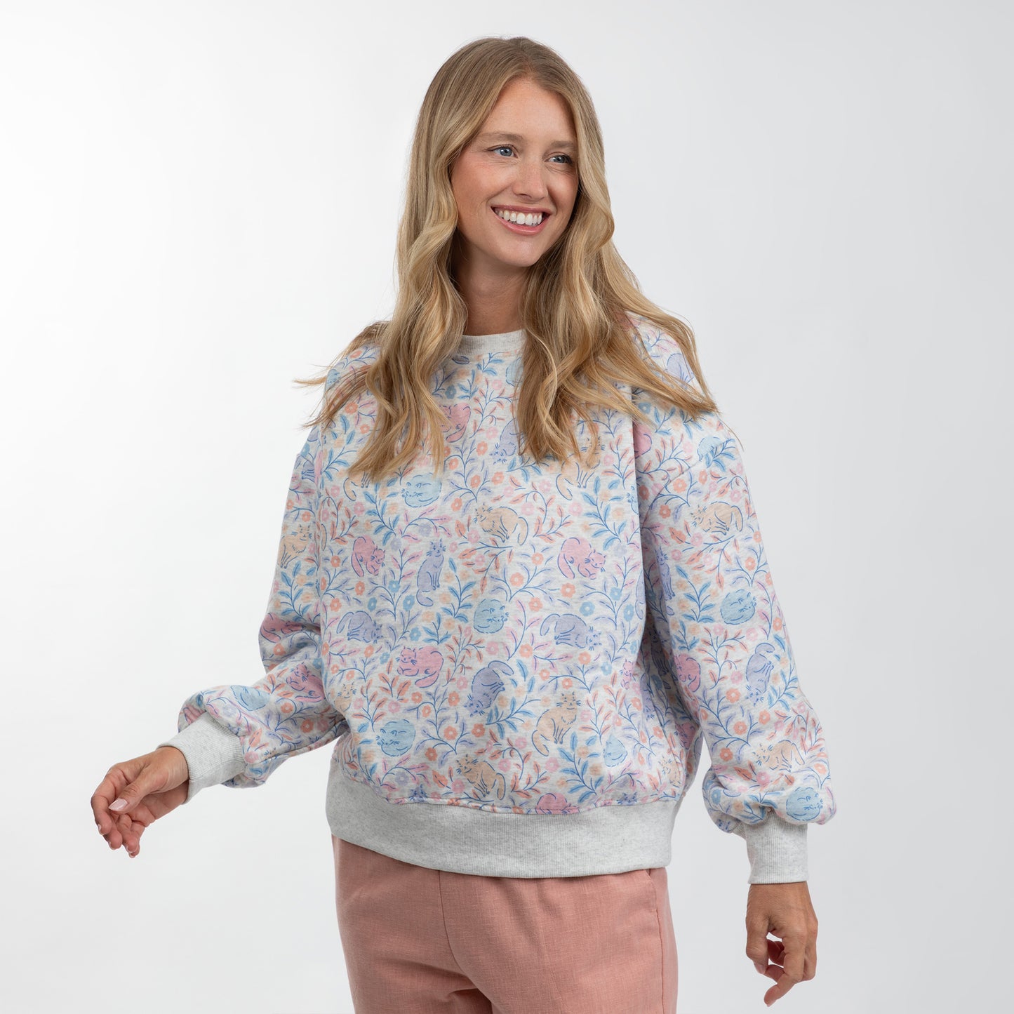 A woman modeling a light gray crewneck sweatshirt with a pastel multicolor floral pattern and ribbed cuffs and hem, paired with dusty pink pants, against a plain white studio background.
