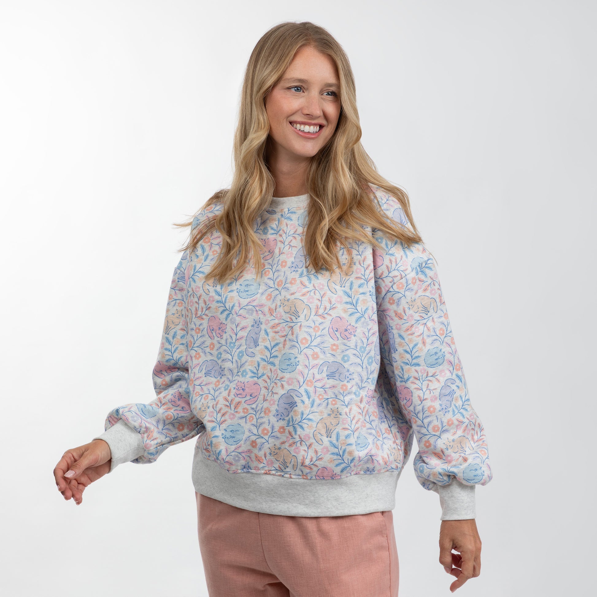 A woman modeling a light gray crewneck sweatshirt with a pastel multicolor floral pattern and ribbed cuffs and hem, paired with dusty pink pants, against a plain white studio background.
