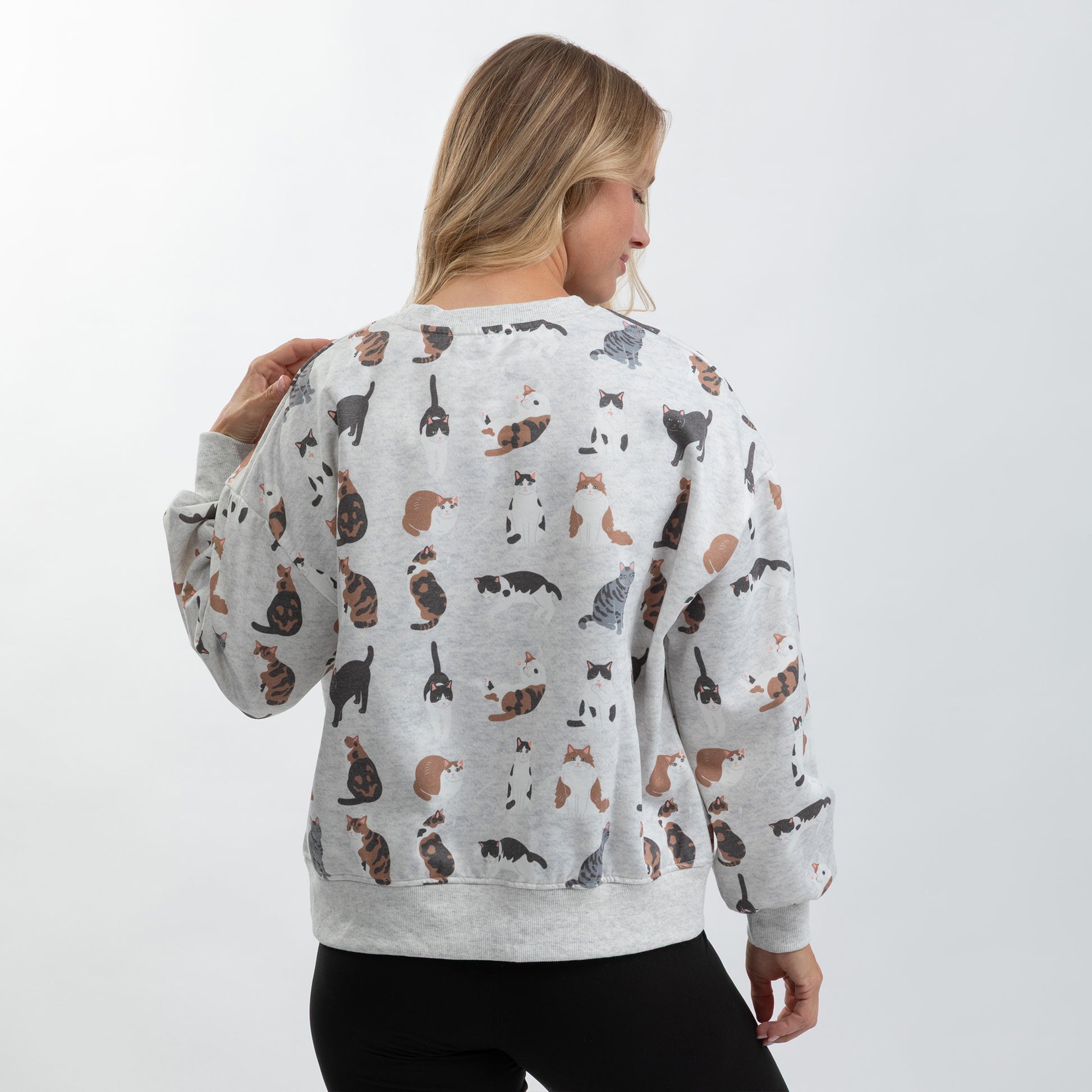 A woman viewed from the back wearing a light gray crewneck sweatshirt covered in a repeating print of various cat illustrations in black, brown, and white, paired with black pants against a plain white studio background.