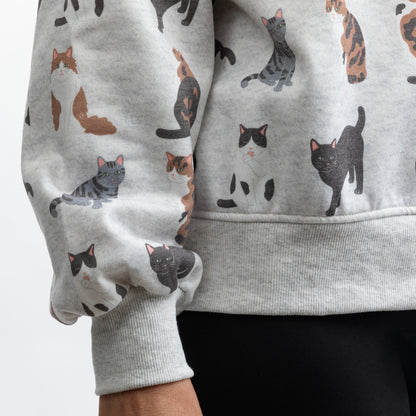 A close-up of a person wearing a light gray sweatshirt with an all-over print of illustrated cats in black, gray, white, and brown. The sweatshirt has ribbed cuffs and a ribbed hem, and is shown paired with black pants against a plain light background.