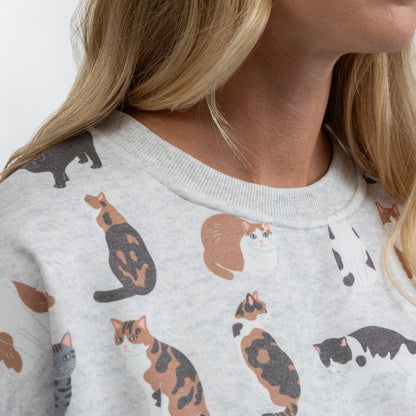 Close-up of a person wearing a light heather gray crewneck sweatshirt with a playful print of various calico and black-and-white cats.