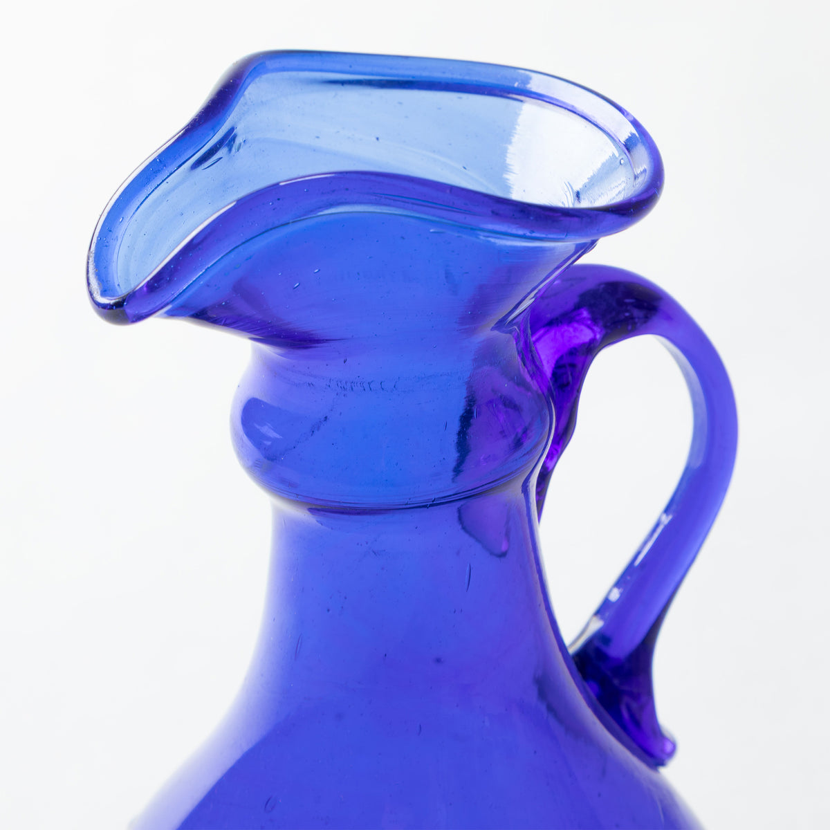 A close-up of a cobalt blue glass pitcher with a wide flared rim and a curved handle, shown against a plain white background.
