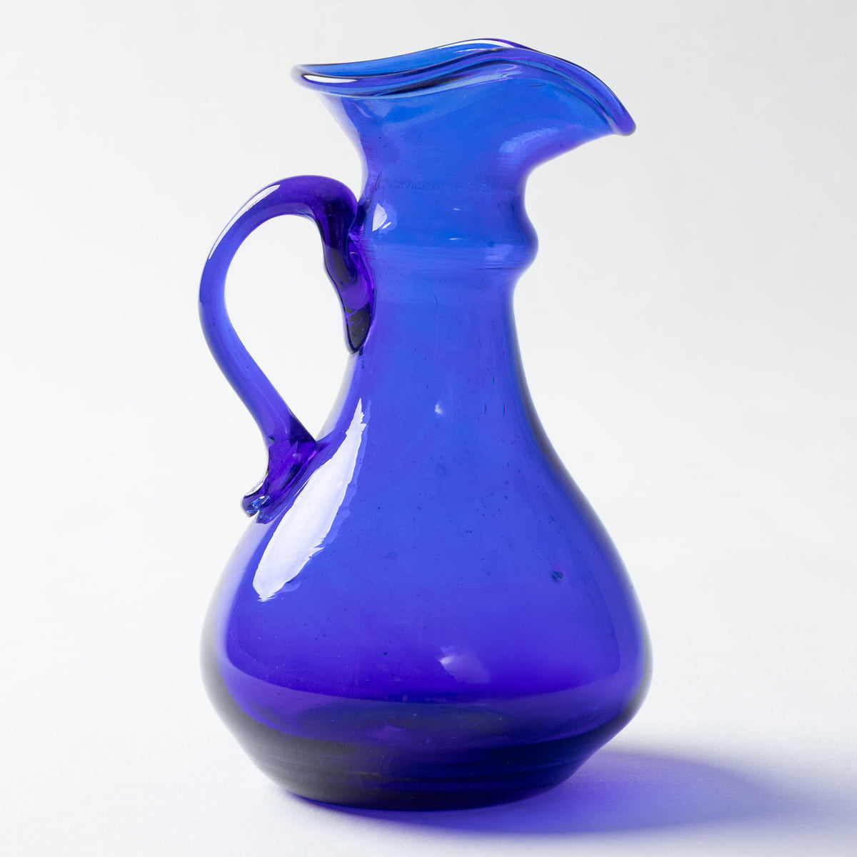 A cobalt blue glass pitcher with a curved handle and a flared spout, shown on a clean white background.