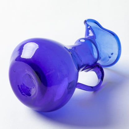 A translucent cobalt blue glass pitcher with a rounded body, small handle, and spout, photographed on a clean white background.