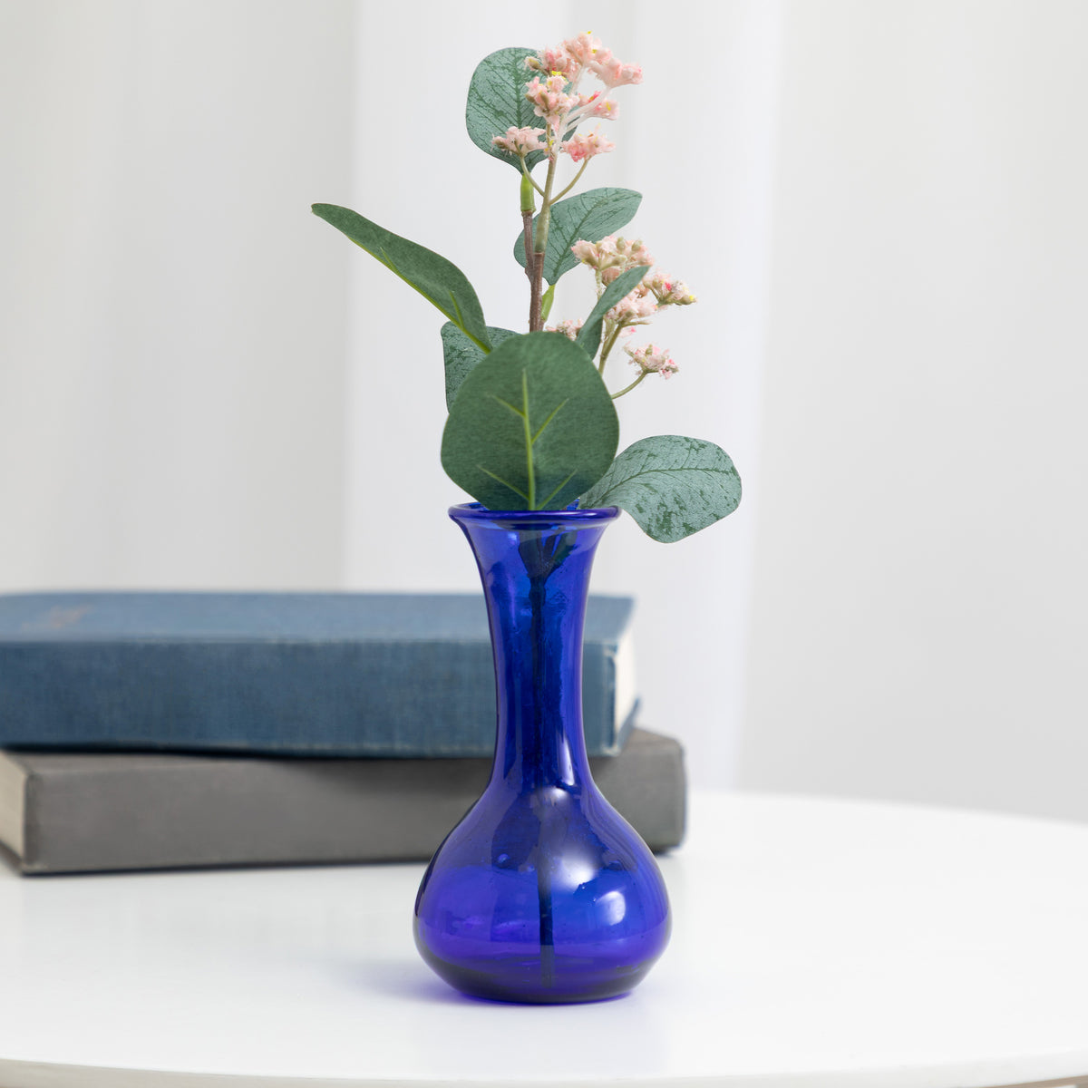 A cobalt blue glass vase with a rounded base, holding a green leafy sprig with small pale pink flowers, set on a white surface with stacked books in the background.