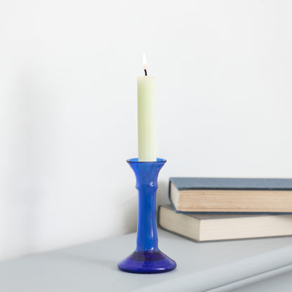 A lit cream-colored pillar candle standing in a cobalt blue glass candlestick holder, placed on a light gray surface with stacked books in the background against a white wall.