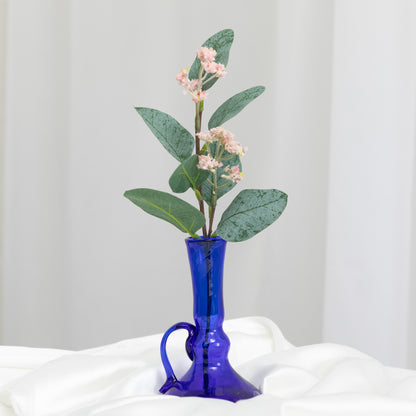 A cobalt blue glass vase with a handle and a curved base, holding a green leafy sprig with small pale pink flowers, set against a soft white draped fabric background.