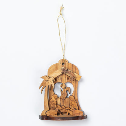 A handcrafted wooden nativity ornament featuring a carved stable scene with a palm tree and the word 'BETHLEHEM' on a light wood background, suspended by a thin string.