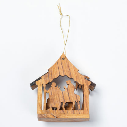 A small wooden hanging nativity ornament with a house-shaped frame and cutout figures, suspended by a thin string on a plain white background.