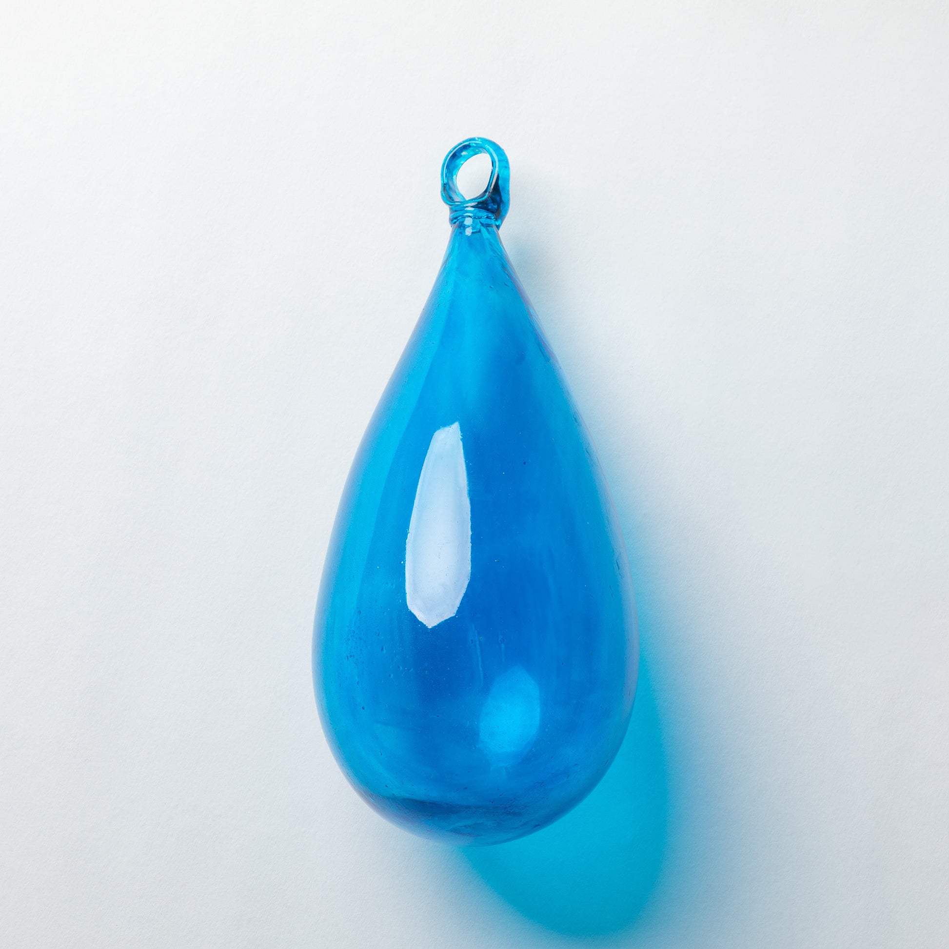A glossy, teardrop-shaped blue glass pendant with a small hanging loop at the top, shown on a clean white background with a soft blue shadow.