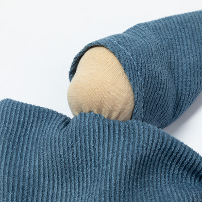 A close-up a blue fabric doll with a wooden pom-pom on the tip of the hat, a wooden pom-pom on the bottom corner, and a small white tag attached.