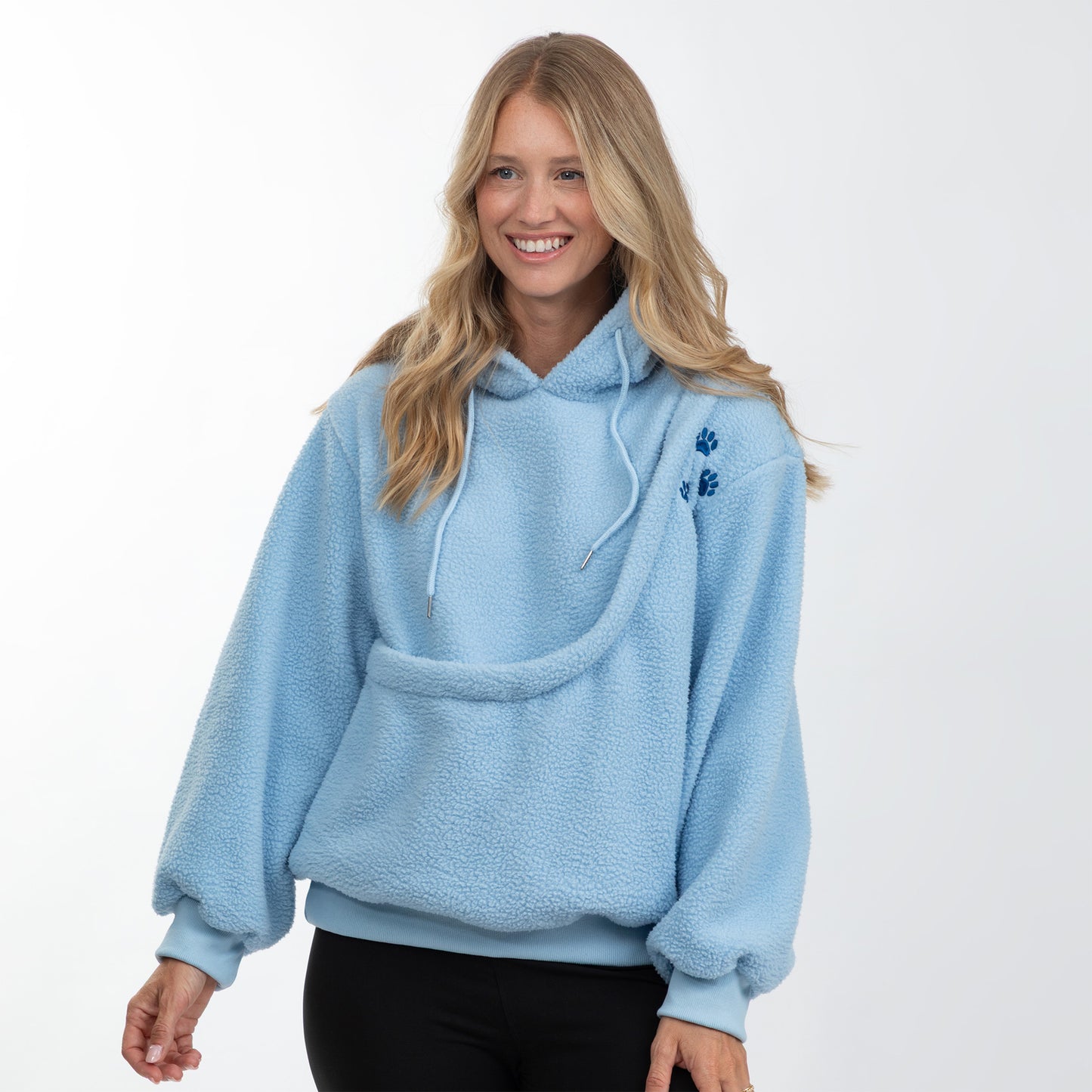 A woman wearing a light blue sherpa fleece hoodie with a large front pouch pocket and small blue embroidered paw prints on the chest, paired with black pants, against a plain white background.