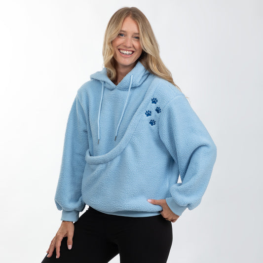 A smiling blonde woman wearing a light blue sherpa fleece hoodie with three dark blue paw prints on the left chest, paired with black pants, against a plain white studio background.