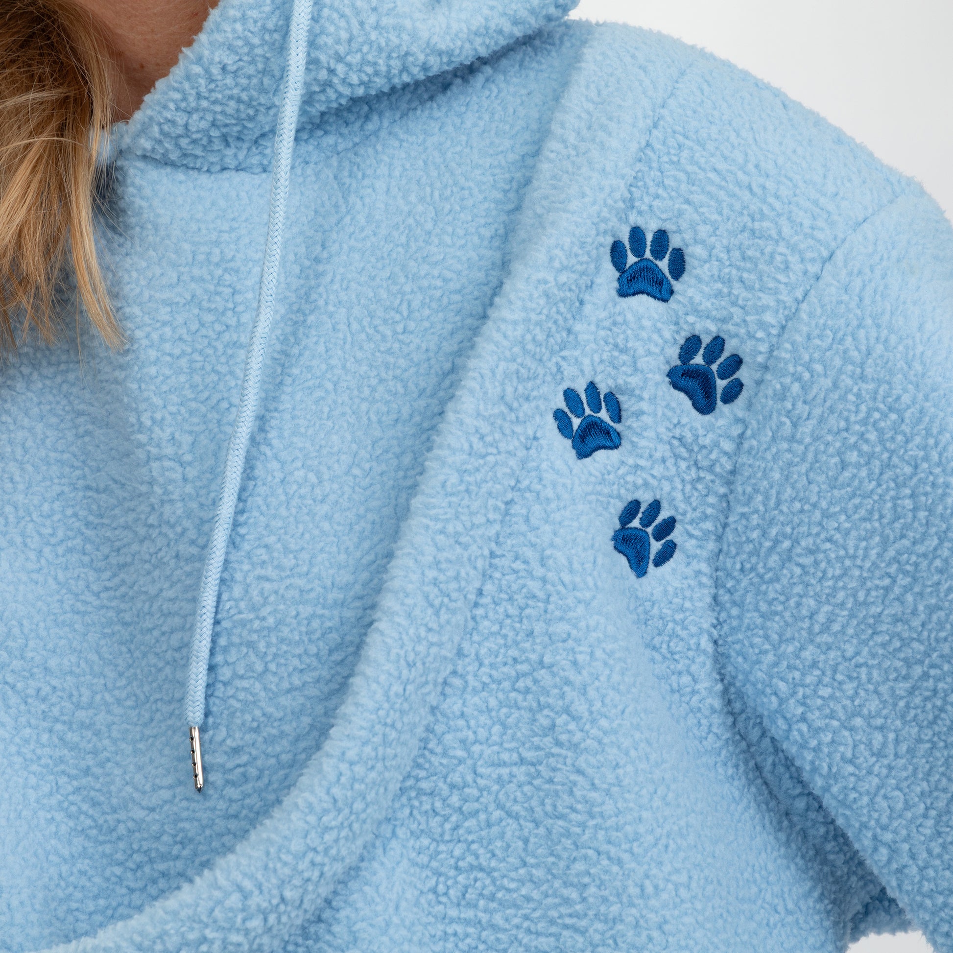 Close-up of a light blue sherpa hoodie with four dark blue paw prints embroidered on the chest and a white drawstring.