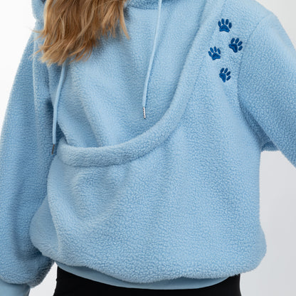 A close-up of a person wearing a light blue sherpa fleece hoodie with a large kangaroo pocket and four dark blue paw prints on the upper left shoulder.