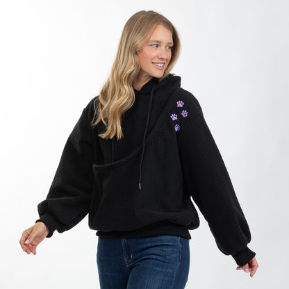 A woman wearing a black fleece pullover hoodie with purple paw print embroidery on the upper left chest, paired with blue jeans, photographed against a clean white studio background.