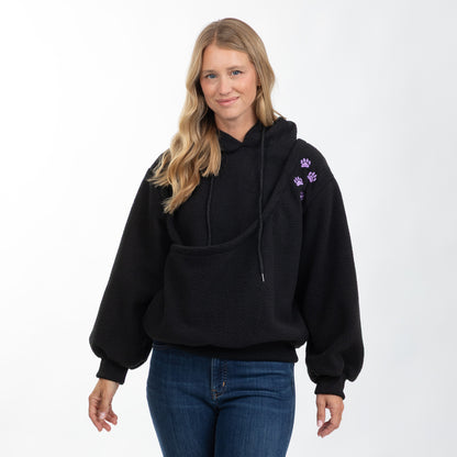 A woman modeling a black fleece pullover hoodie with drawstrings, a front kangaroo pocket, and small purple paw print embroidery on the upper sleeve, paired with blue jeans against a plain white studio background.