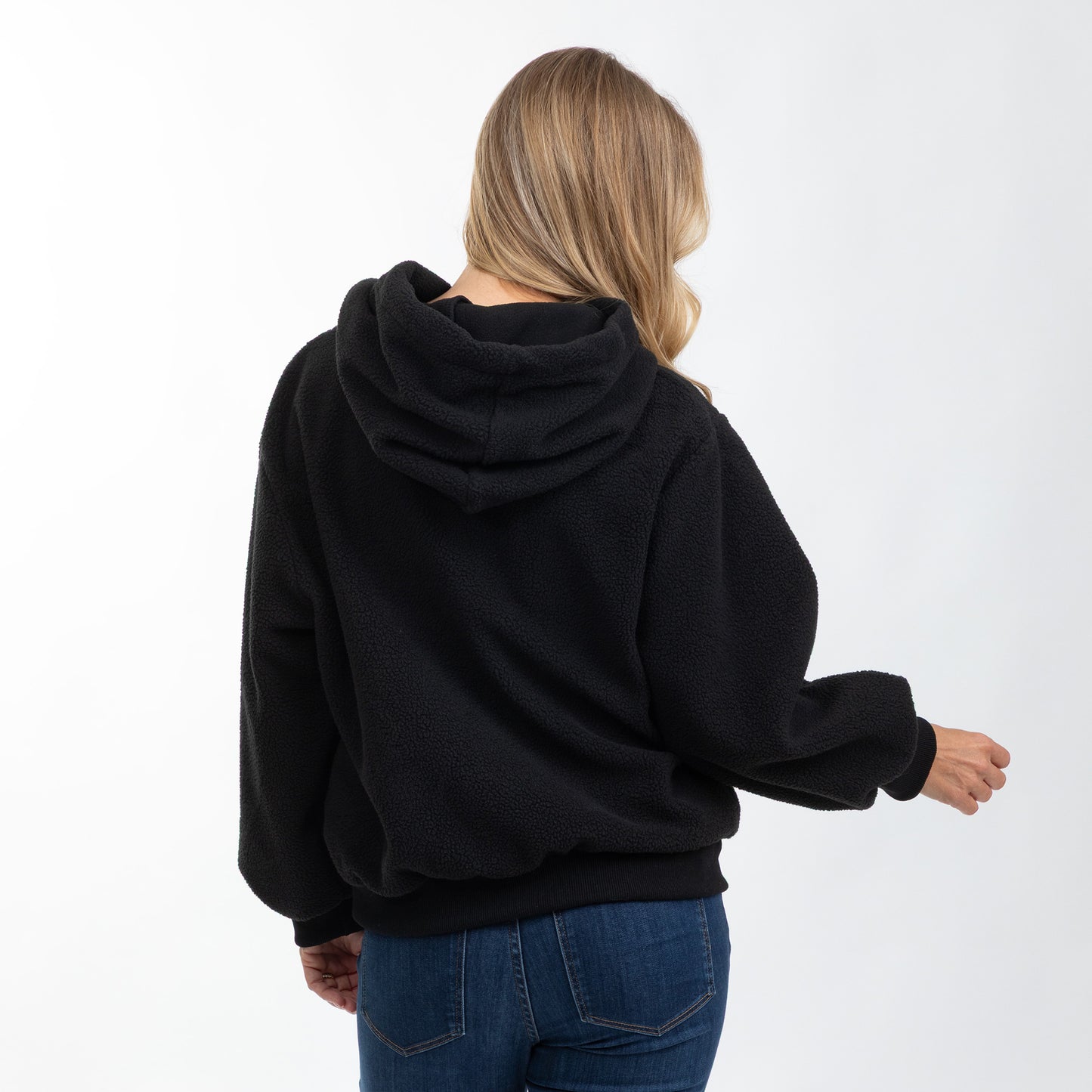 A woman viewed from behind wearing a black fleece pullover hoodie and blue jeans against a plain white studio background.