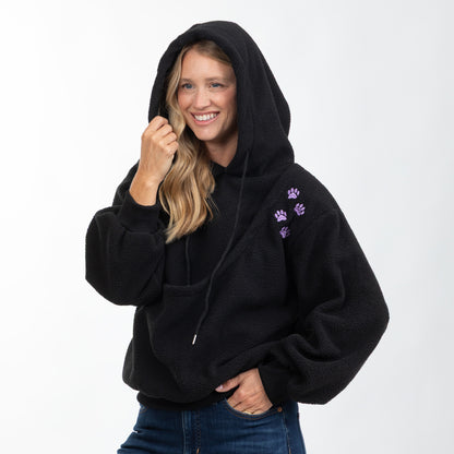 A woman wearing a black fleece hoodie with purple paw print embroidery on the left chest, paired with blue jeans, photographed against a plain white studio background.