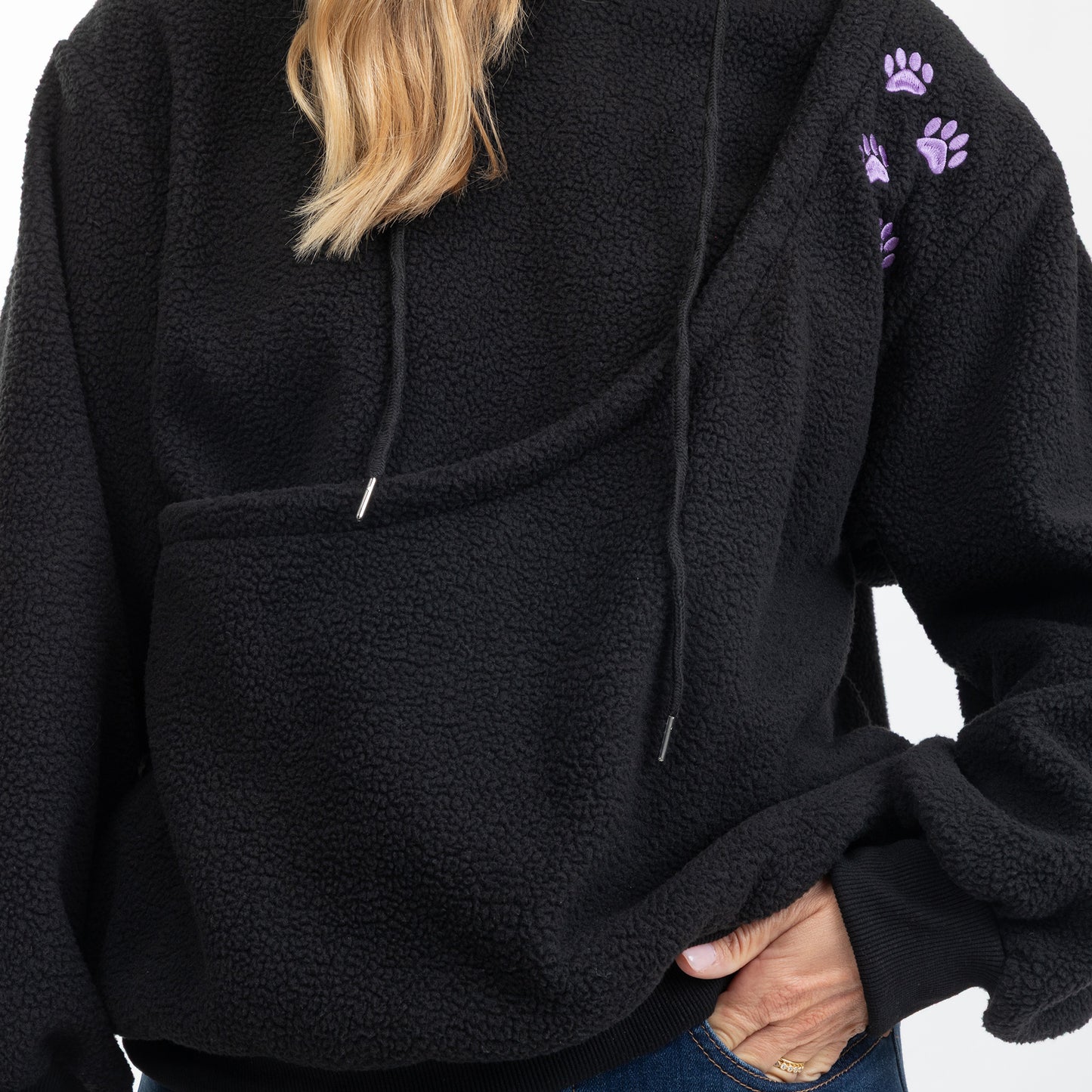 A close-up of a person wearing a black sherpa fleece hoodie with purple paw print embroidery on the upper sleeve, paired with blue jeans.