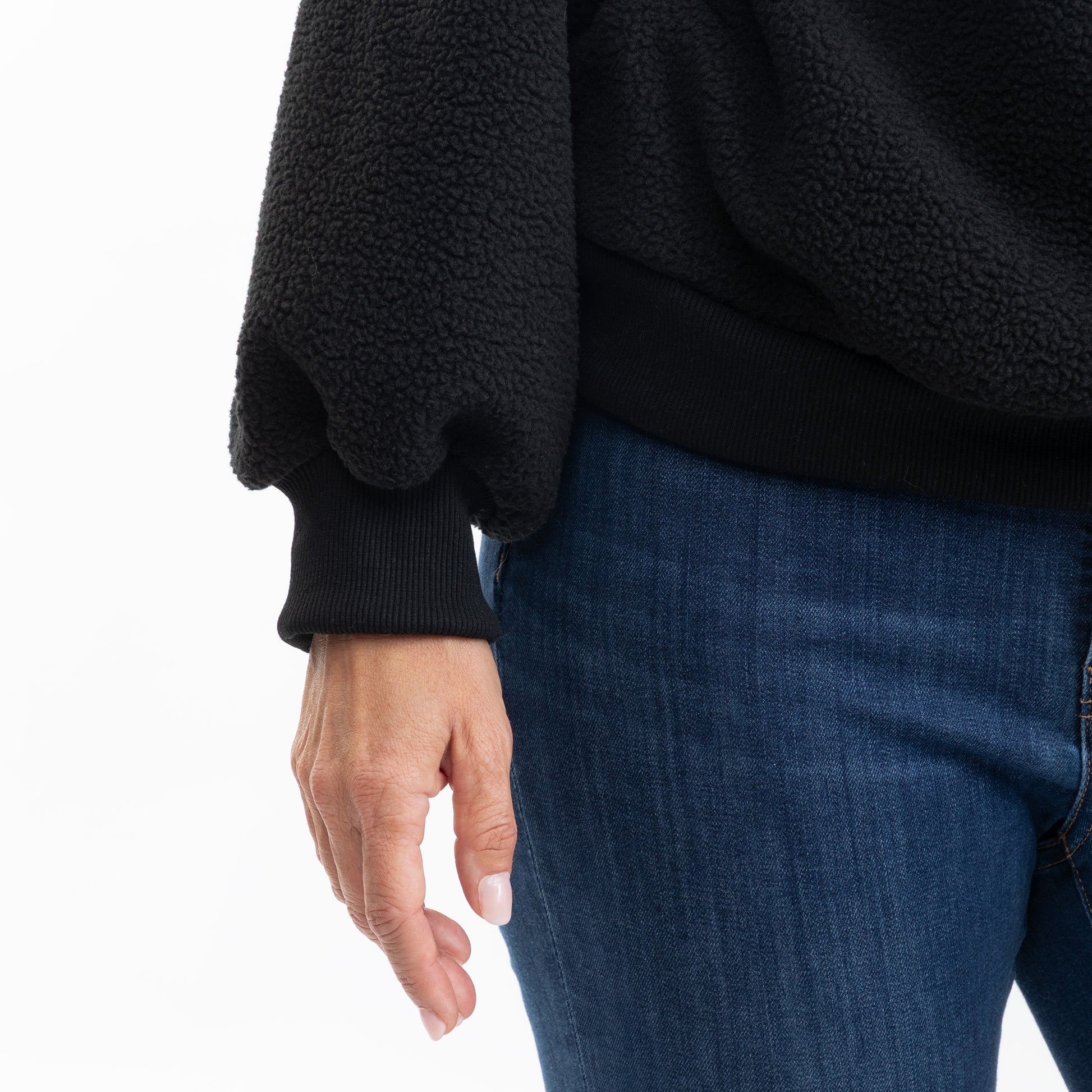 Close-up of a person wearing a black fleece pullover sweater with ribbed cuffs and hem, paired with blue jeans, against a plain white background.