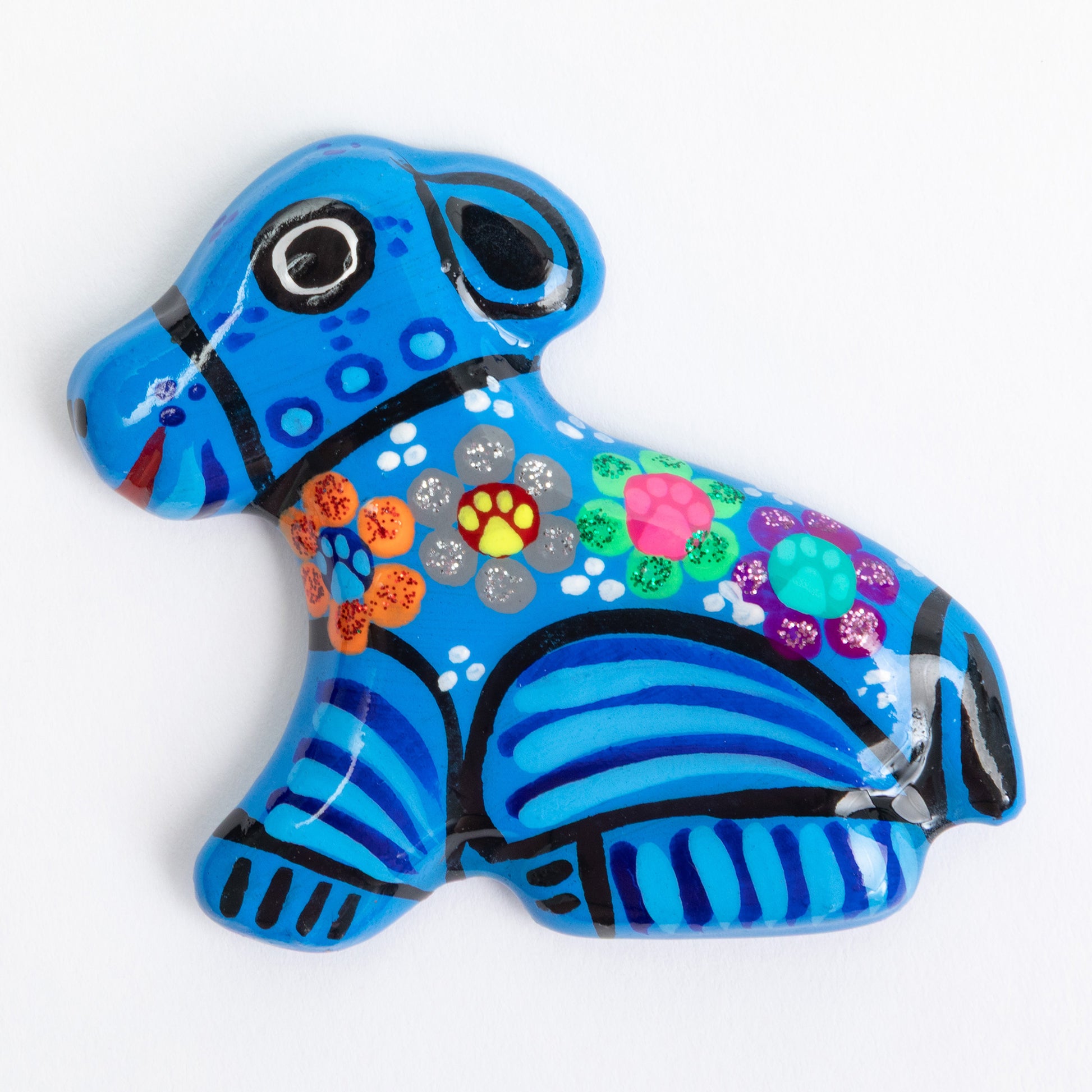 A blue ceramic magnet shaped like a dog, decorated with colorful hand-painted patterns, and black stripes set against a plain white background.