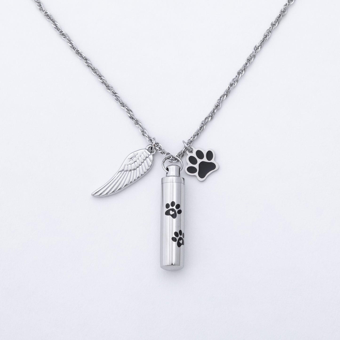 A silver-tone cremation urn necklace featuring a cylindrical vial pendant with black paw print engravings, an angel wing charm, and a black paw-shaped tag on a white background.