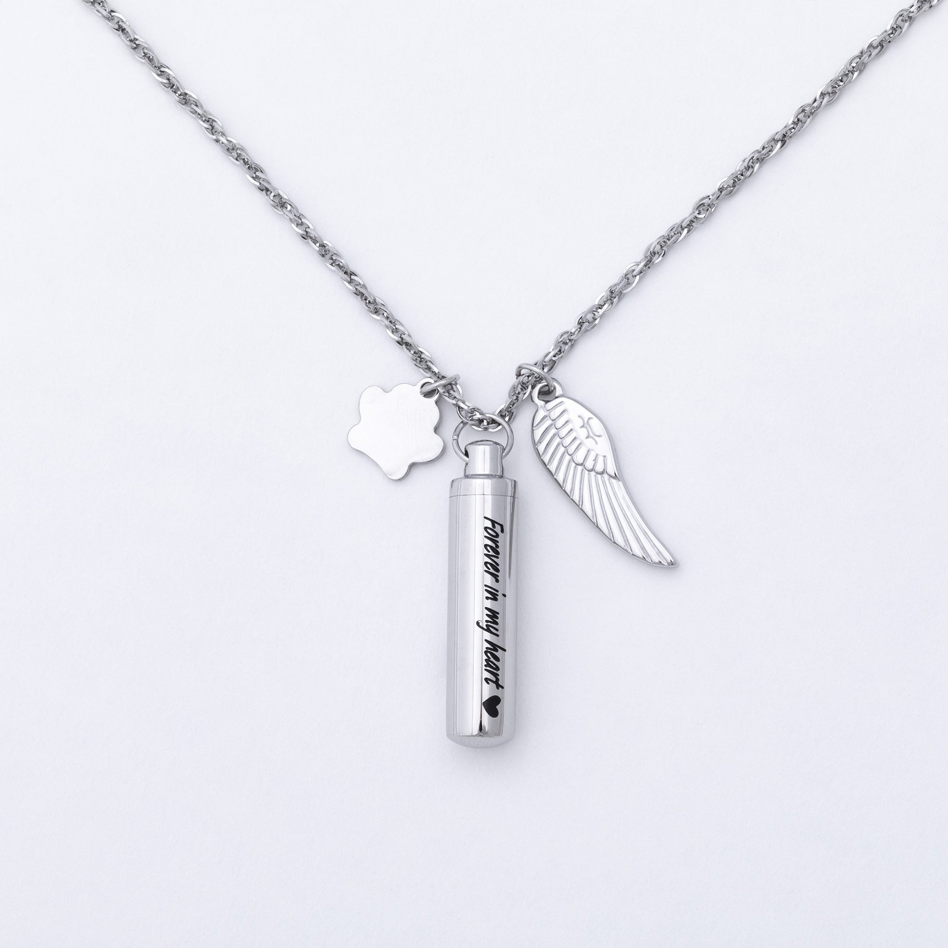 A silver chain necklace featuring three charms: a paw print, a wing, and a cylindrical urn pendant engraved with 'Forever in my heart'.
