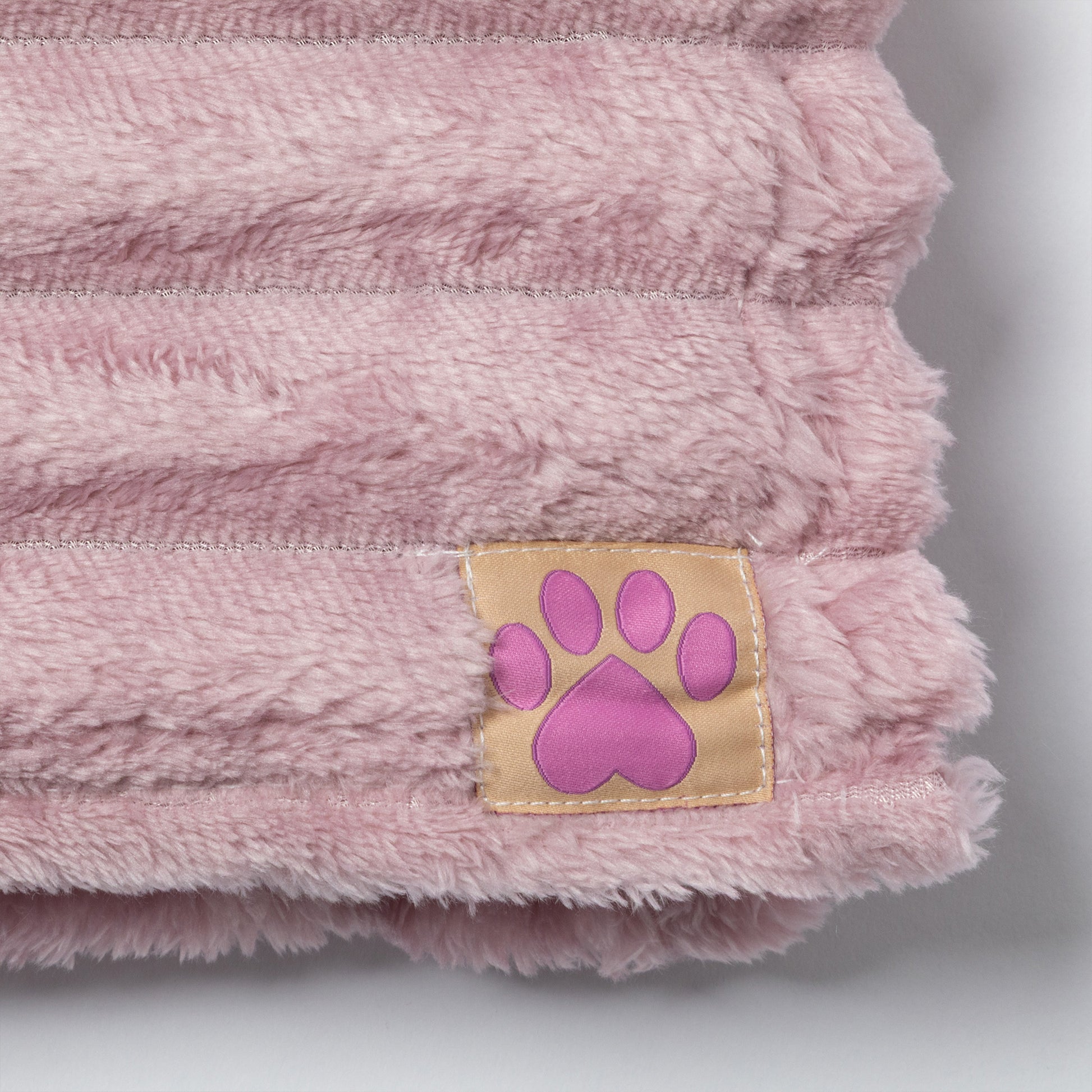 A close-up of a soft pink plush pet blanket with a small tan patch featuring a pink paw print on the corner, set against a neutral gray background.