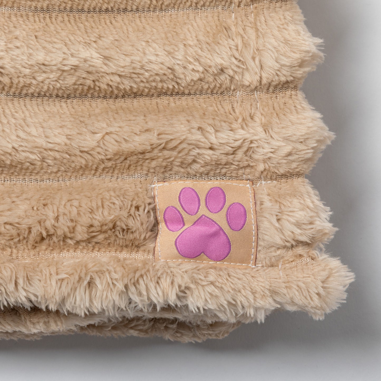 Close-up of a beige plush pet blanket with a small sewn-on patch featuring a pink paw print on a light tan square.