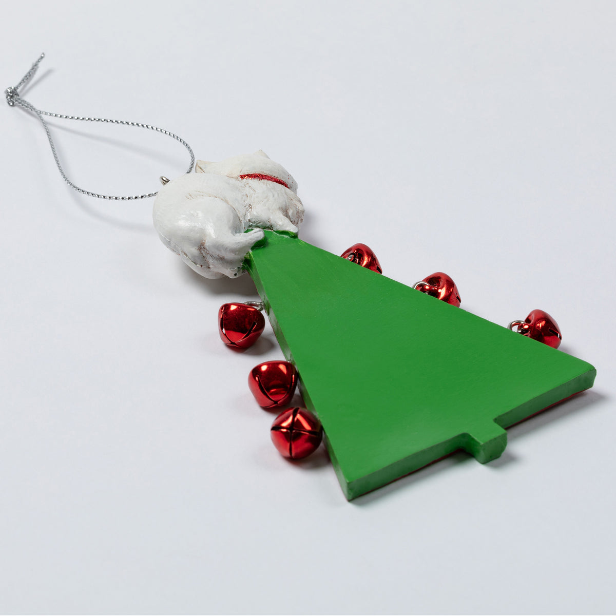 A Christmas ornament shaped like a green Christmas tree with small red jingle bells around it and a white textured figure at the top, hanging from a silver string on a light gray background.