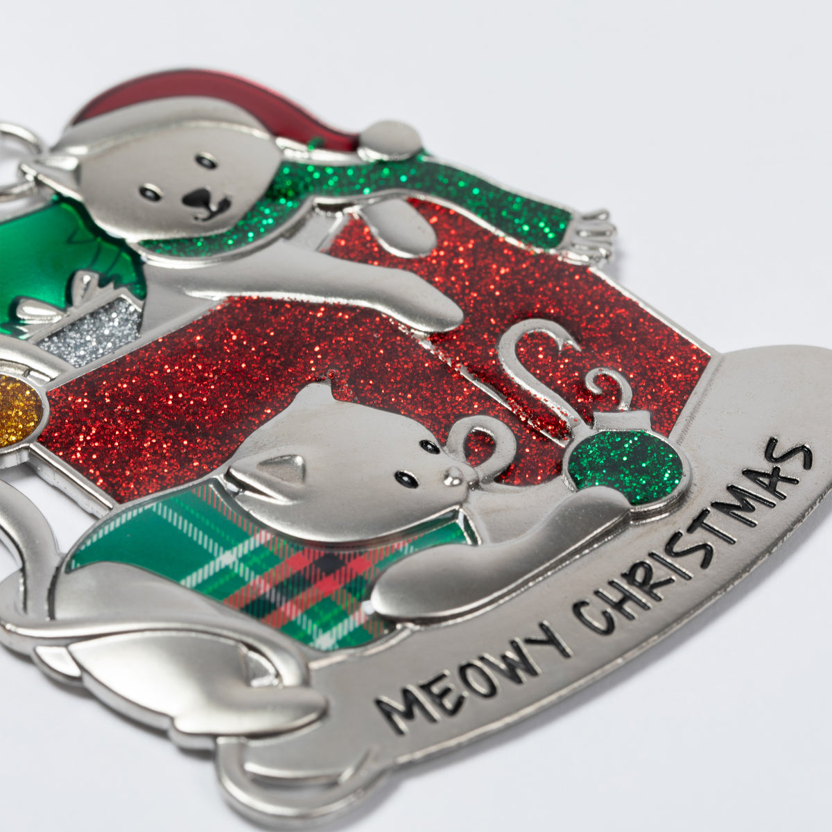 A close-up of a silver metal Christmas ornament featuring a teddy bear and a kitten in holiday attire, with glittery red and green accents and the engraved text 'MEOWY CHRISTMAS' on a white background.