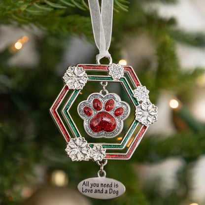 A silver-toned dog paw print Christmas ornament hanging from a white ribbon, featuring red glitter paw print details, a red and green hexagonal frame, and a dangling silver tag that reads 'All you need is Love and a Dog' against a blurred Christmas tree background.