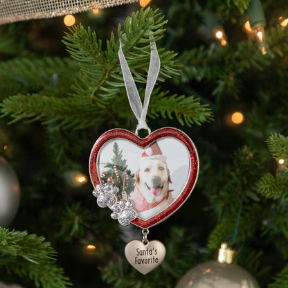 A heart-shaped personalized Christmas ornament hanging on a decorated tree. The ornament has a red glitter border, a photo of a dog wearing a Santa hat, a silver paw print charm, and a dangling heart tag that reads 'Santa's favorite'.