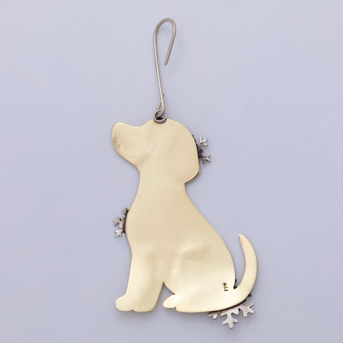 A polished gold-colored metal ornament shaped like a sitting dog, featuring small silver snowflake accents on its ears, paws, tail tip, and bottom edge, hanging from a silver hook against a light lavender background.