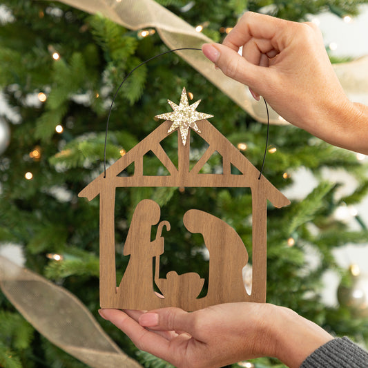 A wooden nativity scene decor held by two hands in front of a decorated Christmas tree. The ornament features a light brown wood cutout of Mary, Joseph, and baby Jesus in a stable, with a glittery gold star on top and a black hanging loop.