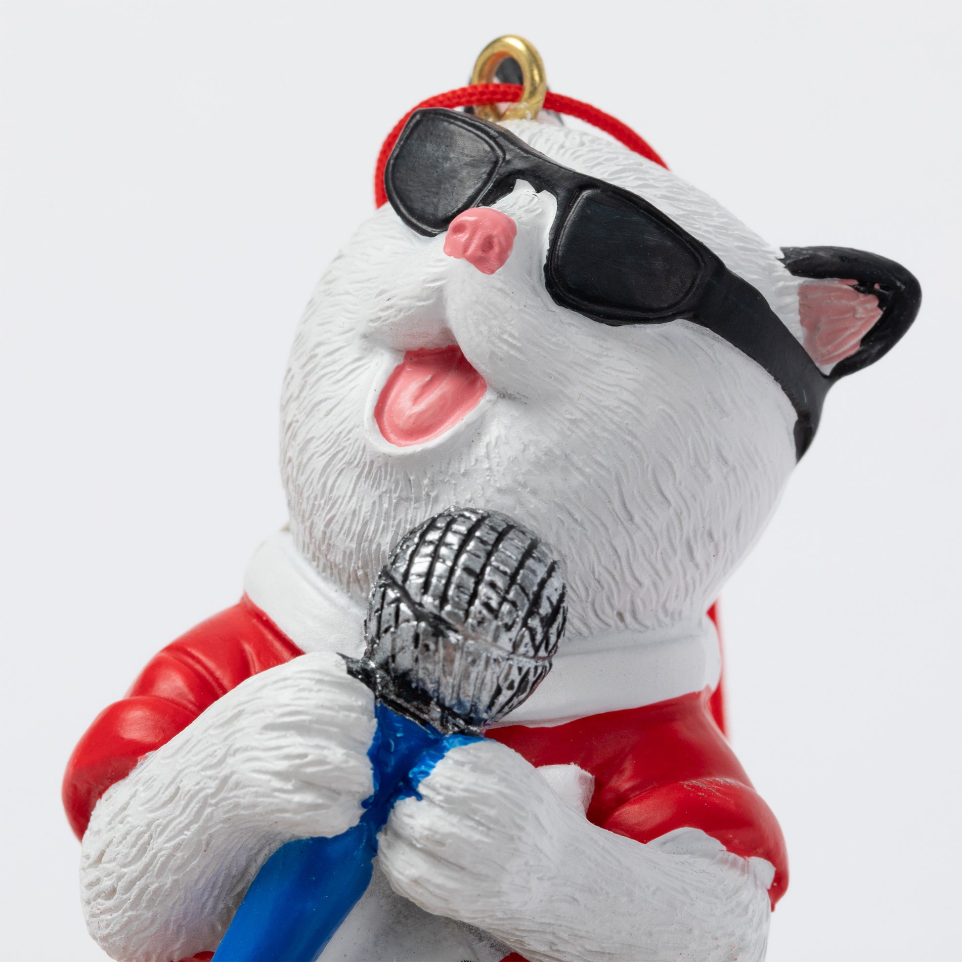 A white cat ornament wearing black sunglasses, a red Santa-style jacket, and a blue bow tie, holding a silver microphone and singing into it. The figurine has a gold hanging loop at the top and is photographed against a plain light gray background.