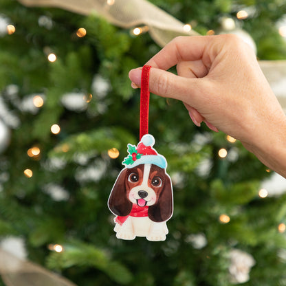 A hand holds a custom dog Christmas ornament shaped like a brown and white Cocker Spaniel wearing a red Santa hat and scarf, set against a blurred Christmas tree background with warm lights.