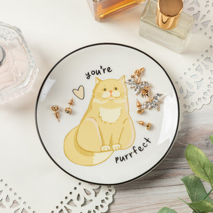 A round white plate with a black rim, featuring a yellow cartoon cat illustration and the words 'you're purrfect'. The plate is styled on a light wooden surface with decorative paper doilies, perfume bottles, and gold and silver earrings placed around it.