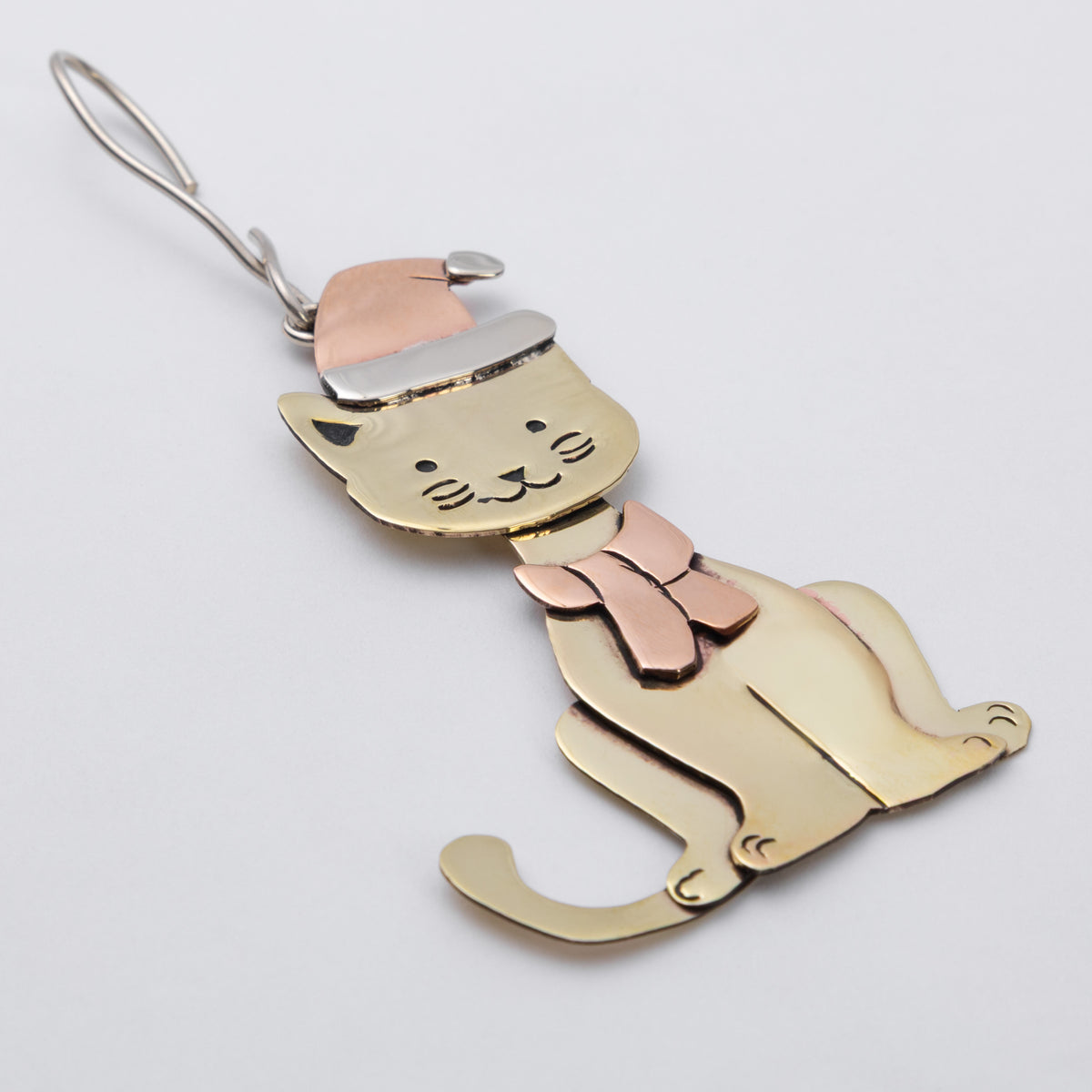 A whimsical cat-shaped ornament with brass body, a copper Santa hat, and matching scarf, hanging from a silver metal hook against a light gray background.