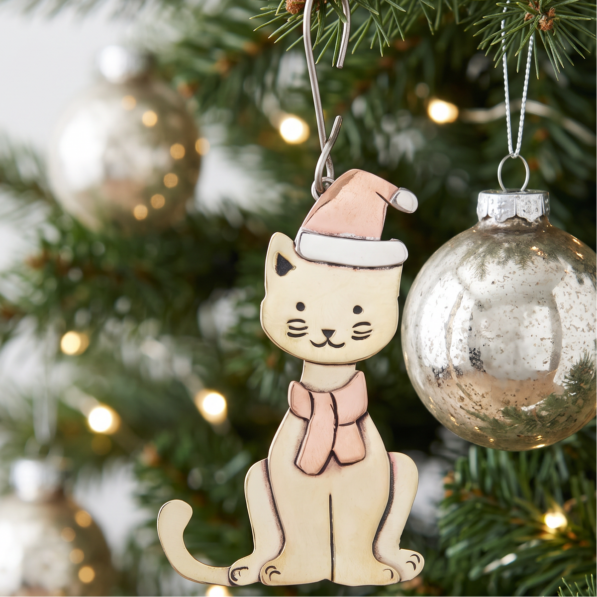 A Christmas tree ornament shaped like a sitting cat wearing a copper Santa hat and matching scarf, hanging from a silver wire hook among green pine branches with blurred lights and reflective ornaments in the background.