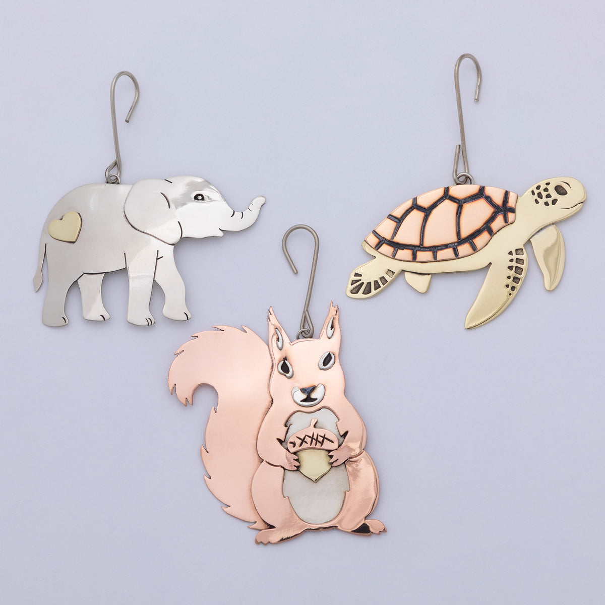 Three metal animal-shaped wall hooks or ornaments hanging against a light lavender background: a silver elephant with a gold heart patch, a cream turtle with dark linework, and a peach squirrel holding a gold acorn.