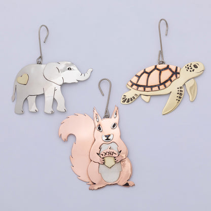 Three metal animal-shaped wall hooks or ornaments hanging against a light lavender background: a silver elephant with a gold heart patch, a cream turtle with dark linework, and a peach squirrel holding a gold acorn.