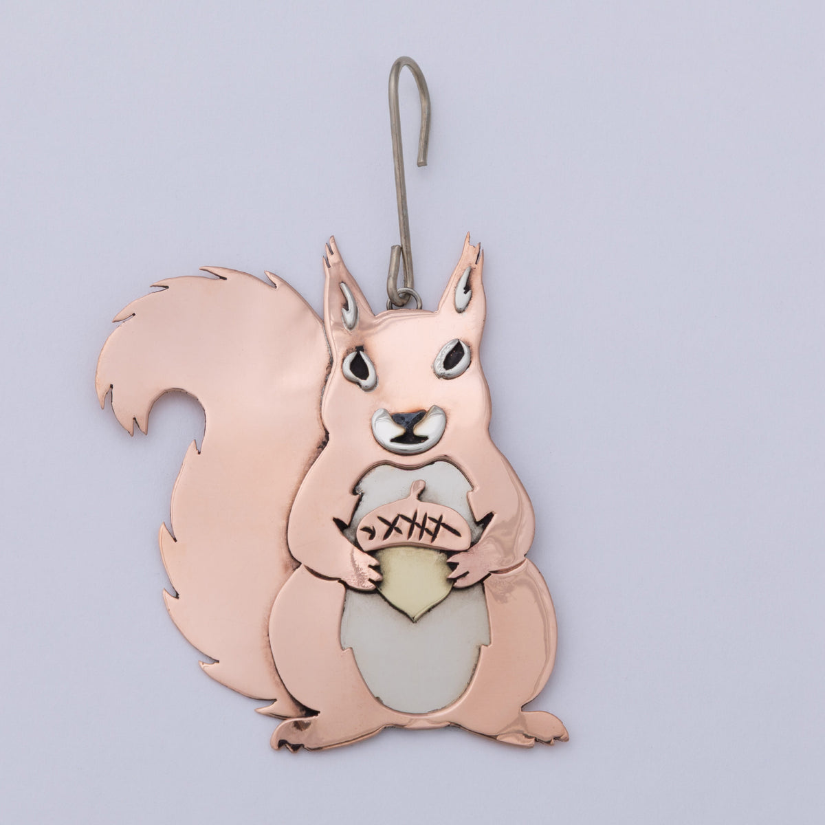 A copper-colored metal squirrel ornament holding an acorn, with a silver hanging hook at the top, shown against a light lavender background.