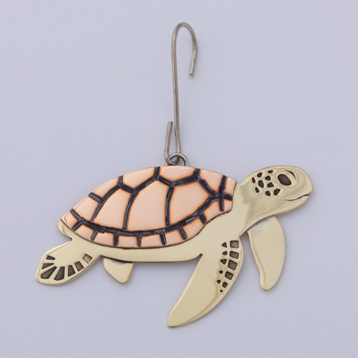 A sea turtle ornament with a gold-toned body and a peach-colored shell outlined in black, hanging from a silver metal hook against a light gray background.