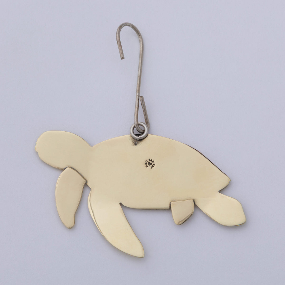 A sea turtle-shaped metal ornament in a warm gold finish, suspended from a silver-colored metal hook with a small decorative emblem on its shell, set against a plain light gray background.