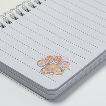 A close-up of a spiral-bound lined notebook with a small colorful floral sticker or illustration near the bottom right of the page, photographed on a clean white background.