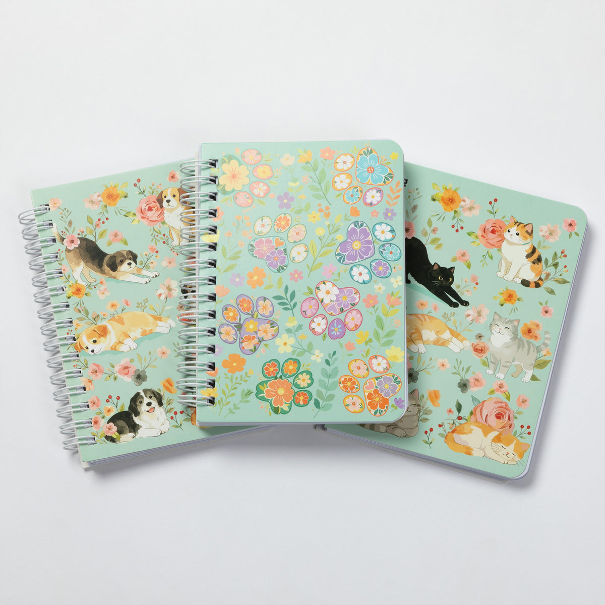 Three spiral-bound notebooks with pastel floral and cat illustrations fanned out on a light gray background.