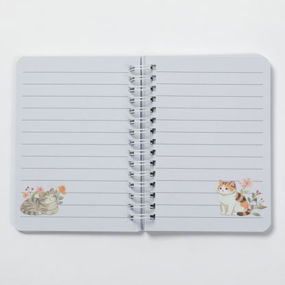 An open spiral-bound lined notebook with two watercolor cat illustrations decorated on the left and right pages, set against a clean white background.